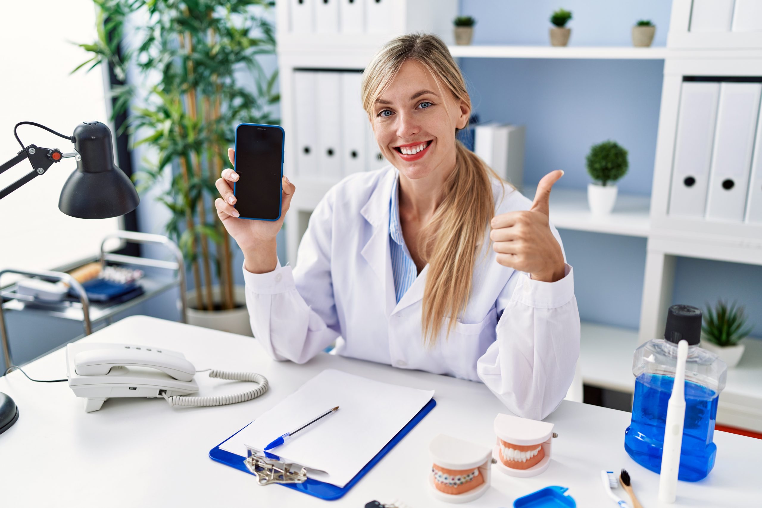 Uma dentista sorrindo para a câmera, sentada em sua mesa do consultório, segurando um smartphone e fazendo sinal positivo com o dedo.